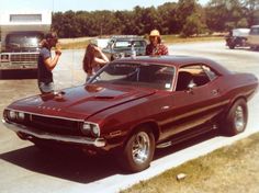 two people standing next to a red muscle car