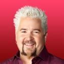 a man with white hair and beard wearing a purple shirt smiles at the camera while standing in front of a red background