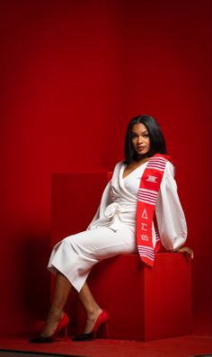 a woman sitting on top of a red block wearing a white dress and a red sash