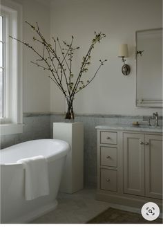 a white bath tub sitting under a window next to a sink and a vase with flowers