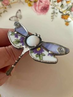 a hand holding a glass and metal dragon brooch with flowers on it's wings