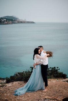 a man and woman embracing each other on top of a hill next to the ocean