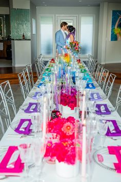 a couple standing next to each other in front of a long table with flowers on it