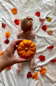 a person holding a small pumpkin in front of a baby on a bed with leaves