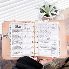 a hand holding an open planner book on top of a wooden table next to a potted plant