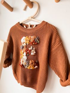 an orange sweater with flowers on it hanging from a wooden hanger next to a white wall