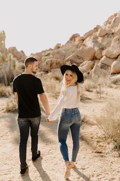 a man and woman holding hands walking through the desert with rocks in the back ground