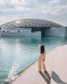 a woman standing on the edge of a body of water near a building with a domed roof