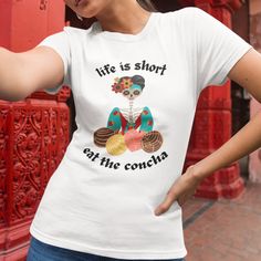 Life is Short Eat the Concha T-shirt is a vibrant and playful Hispanic-themed t-shirt that celebrates the joy of life and the deliciousness of traditional Mexican pastry, the concha. The t-shirt features a bold and colorful design with an image of conchas and a 'sweet' Vieja offering you these delicious pan dulces. This humorous and light-hearted t-shirt is perfect for those who appreciate Hispanic culture, love Mexican cuisine, or simply want to add a touch of fiesta to their wardrobe. Whether worn at a party, a cultural event, or just for everyday fun, this t-shirt is sure to spark conversations and smiles wherever you go! Viva la vida y viva la concha! All of our items are created when ordered, typically done within 3 business days, then out for shipping.  (My daily order cut off is 1pm White Novelty Crew Neck Top, Novelty White Crew Neck Top, White Novelty Cotton T-shirt, White Cotton Novelty T-shirt, White Novelty Crew Neck T-shirt, White Novelty Tops As Gift, White Novelty Tops For Gifts, White Novelty Short Sleeve Top, Novelty White Short Sleeve Top