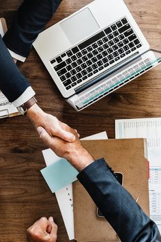 two people shaking hands in front of a laptop on a wooden table with papers and other office supplies