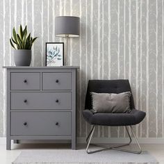 a grey chair next to a dresser with a lamp on it and a plant in the corner