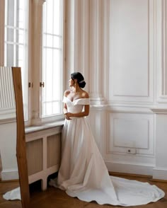 a woman standing in front of a window wearing a wedding dress and looking out the window