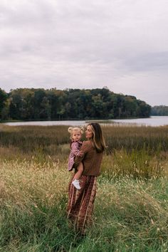 a woman holding a small child in her arms while standing in tall grass near a body of water