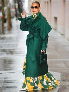 a woman in a green coat and yellow dress is walking down the street with her hand up