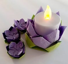 a white candle sitting on top of purple paper with flowers next to it and one light in the middle