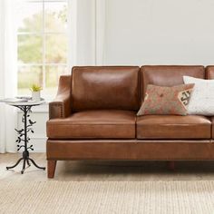 a brown leather couch with two pillows on it in front of a white wall and window