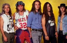 four men standing next to each other in front of a yellow wall with long hair