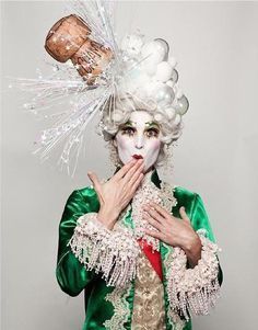 a woman with white makeup and green dress is holding a bottle in her hand while wearing an elaborate hat