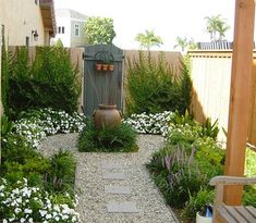 a small garden with white flowers and plants around the water fountain, along with a wooden bench