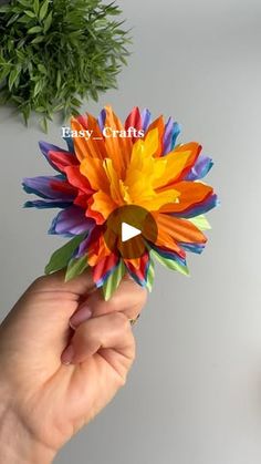 a hand holding a colorful paper flower in front of a potted plant