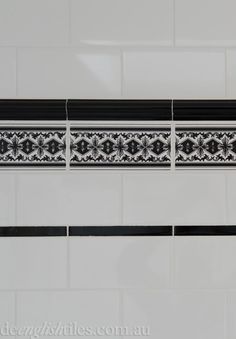 a white tiled wall with black and white designs on the border, in a bathroom