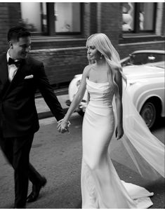 a man and woman holding hands walking down the street in front of a white car