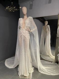 a white dress with long sleeves and cape on display in front of two mannequins