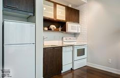 an empty kitchen with white appliances and wood flooring in front of a brick wall