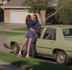 two people kissing in front of a green car on the side of the road next to a house