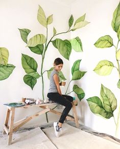 a woman sitting on top of a wooden bench next to a green plant painted on the wall