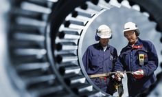 two men in hardhats are standing next to an industrial gear wheel and looking at the camera