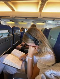 a woman sitting in an airplane reading a book and wearing headphones on her ears