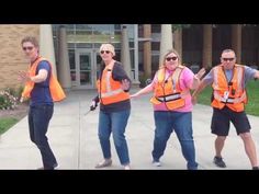 four people in orange vests are dancing on the sidewalk with their arms out and one person is wearing sunglasses