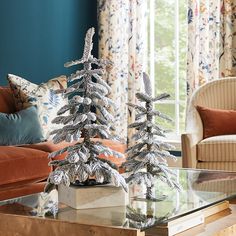 two silver christmas trees sitting on top of a glass table in front of a couch