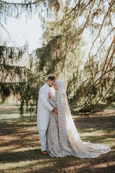 a bride and groom standing in front of a large tree with their arms around each other