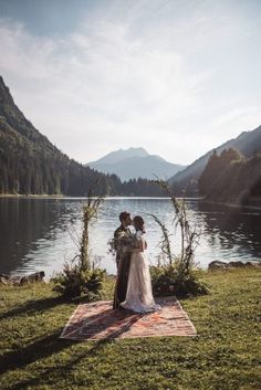 a bride and groom standing in front of a lake surrounded by greenery with mountains in the background