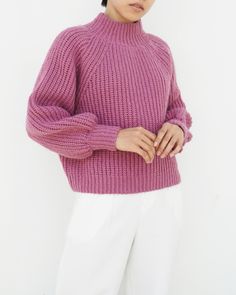 a woman wearing a pink sweater and white pants with her hands on her hips, standing in front of a white wall