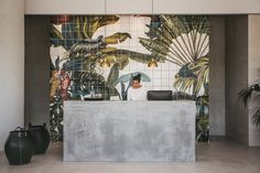 a woman sitting at a counter in front of a tiled wall with palm leaves on it