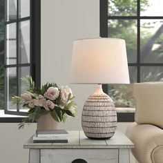 a table with a lamp and flowers on it next to a couch in front of a window