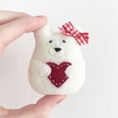 a hand holding a small white teddy bear with a red heart