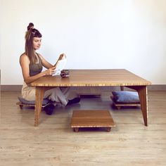 a woman sitting at a wooden table with a book in her hand and a dog laying on the floor