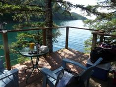 two chairs and a table on a deck overlooking the water with pine trees in the foreground