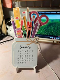 a desk calendar with scissors and pens in it on top of a table next to a computer