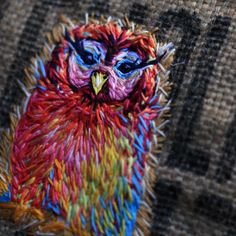 an embroidered owl sitting on top of a piece of fabric with blue eyes and multicolored feathers