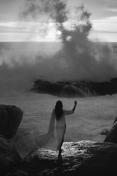 a woman standing on top of a rock next to the ocean under a large wave