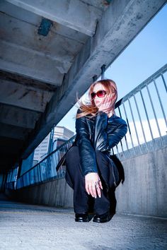 a woman sitting on the ground with her hands in her mouth and wearing sunglasses, leaning against an overpass