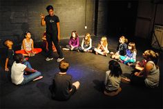 a group of people sitting on the floor in a circle with one person standing up