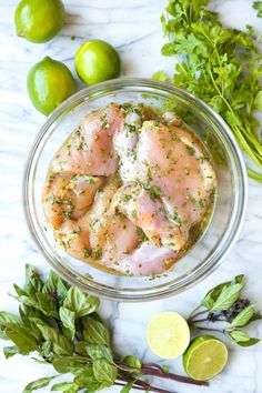 raw chicken breast in a glass bowl surrounded by fresh herbs and limes on a marble countertop