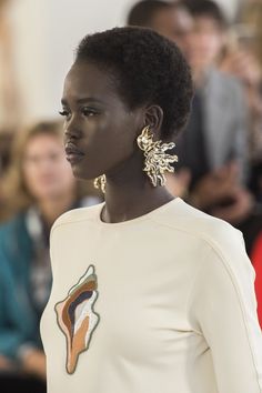 a woman with short hair wearing gold earrings and a white dress in front of a crowd