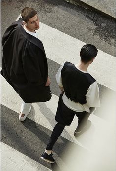 two people walking across a crosswalk in the middle of the street, one is wearing a black and white outfit